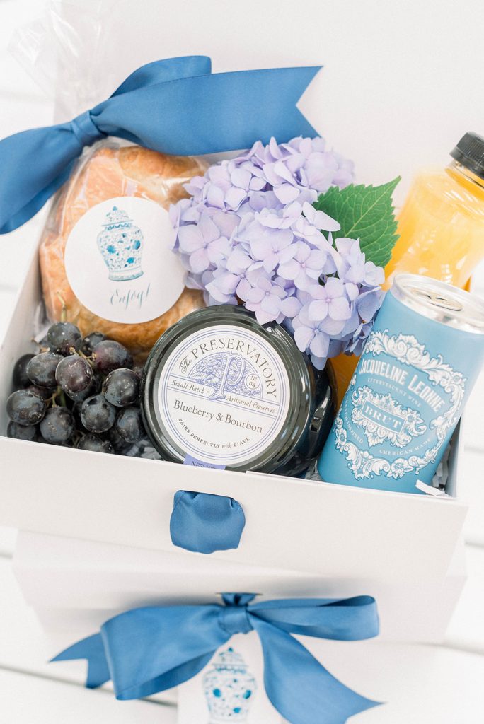 A brunch-themed wedding welcome gift designed by The Welcoming District. These pretty gifts welcomed guests at Catesby Farm Estate in Virginia. Gift goodies included a croissant, grapes, fresh orange juice, champagne, and blueberry preserves. Each was finished with blue satin ribbon and ginger jar gift tags and labels.